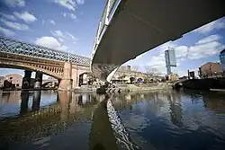 A photo taken from underneath a modern bridge over a river, with the underside of the bridge curving from the top right to the centre to the centre right of the frame. In the background is a grand railway bridge approximately perpendicular to the viewer which the first bridge then becomes parallel to the right of, and there is a skyscraper on the centre right as well.