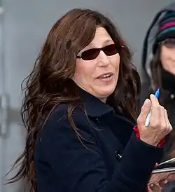 Image of Catherine Keener's head and torso. She is wearing a heavy coat and dark sunglasses. Keener is holding a blue Sharpie and appears to be signing autographs.