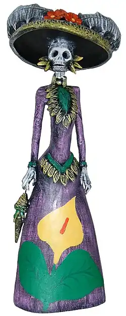 A photograph of a sculpture of a skeletal figure in a brightly colored, feminine outfit and hat.