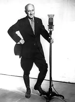 Full body photograph of DeMille wearing a blakc suit, holding a top hat in one hand and the CBS radio microphone in the other