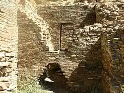 Inside daytime view of a ruined and ceiling-less rectangular room. Tawny-beige stacked sandstone bricks compose walls rising from brush-covered ground. The several walls visible in the image are up to perhaps a dozen feet in height. In the wall immediately at center, a triangle-shaped entrance several feet high leads to an adjacent chamber behind. The upper part of the same wall, shaped like an inverted-triangle, has fallen away or otherwise been removed, revealing a rectangular doorway leading to yet another concealed room. At left and right are two similar walls perpendicular to the one at middle.