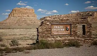 Large circular depression outlined by a stone wall. The bottom is flat and grassy, and has a collection of rectangular stone foundations and smaller circles of stone. A great sandstone cliff towers in the background, and beneath the cliff are other stone foundations that are larger and higher.