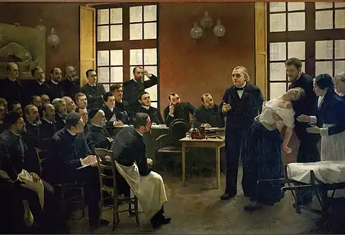 A painting of a 19th-century medical lecture. At the front of the class, a woman faints into the arms of a man standing behind her, as another woman, apparently a nurse, reaches to help. An older man, the professor, stands beside her and gestures as if making a point. Two dozen male students watch them.
