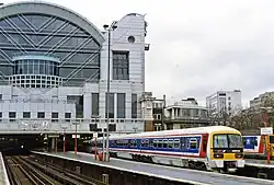 An NSE-branded Class 465, a commuter train, at a very grand terminus station. The skyline of London is visible in the background and about two-thirds of the station building. The photo is taken from quite a few platforms away