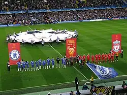The Chelsea (blue) and Liverpool (red) teams line up side-by-side in front of banners bearing their club crests and the Champions League final logo. In the background, a flag bearing the Champions League logo is waved over the centre circle, while the main stand is further back, full of spectators. In the foreground, Chelsea fans wave blue flags bearing their club crest.