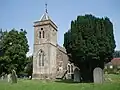 Gray stone building. Prominent square tower with arched window, topped by small slate pyramidal roof. Left and right of the building are yew trees amongst gravestones.