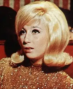 Cher with a voluminous blonde bouffant hairstyle and winged eyeliner, wearing a gold sequined top while seated in a red booth.