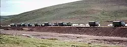 Line of military trucks parked along a road in a sparsely populated mountainous landscape, with grassy hills in the background and temporary structures visible beside the road.
