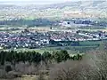 Multiple houses mostly with red roofs seen in a valley between the vegetation in the foreground and the hills beyond.