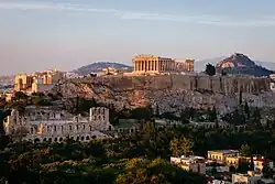 Color photograph of a cityscape, with a slightly forested landscape in the foreground, a rock face in the middle and rolling hills in the background. Ruins of large Classical buildings can be seen in the foreground and middle,