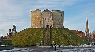 A photograph of a small castle on top of a green mound; the castle has three circular walls visible. Behind the castle the sky is overcast and dark grey.