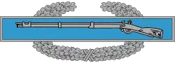 A metal device depicting a blue bar with a rifle, in front of a wreath of silver leaves.