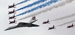 BA-liveried Concorde leading a V-formation with seven of the Red Arrow's Gnat aircraft in view. The Gnats are trailing blue and white smoke