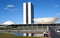 The view shows a 20th-century building with two identical towers very close to each other rising from a low building which has a dome at one end, and an inverted dome, like a saucer, at the other.