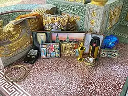Contemporary joss paper, Thailand, consisting of paper T-shirts, TV, mobile phone, electric fan, radio, electric kettle, tea set, oil lamp, watches, glasses, necklaces, shoes, and other accessories, along with a bag of traditional joss paper and bamboo rings marking the burning area. Shoes are usually burnt last.