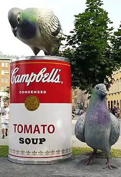 a sculpture of a red and white Campbell's Tomato Soup can with a bird to its right and a bird perched atop it looking down at it. It seems to be situated in front of a municipal area with buildings and people in the background.