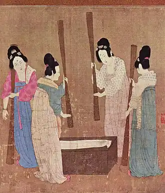 A group of women wearing high-waisted skirts, wrap-front tops and large hair buns use wooden rods to prepare a length of white silk.
