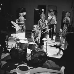 The band Cream is shown playing on a TV show. From left to right are drummer Ginger Baker (sitting behind a drumkit with two bass drums) and two electric guitarists.