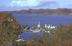 A small harbour surrounded by trees offers shelter to several yachts, fishing boats and a motor launch. The harbour is connected to the sea beyond by a lock gate. A small skerry lies in the water and brown rocky hills dominate the distance.