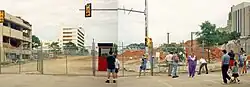 Two images are stitched together showing the site of where the building stood prior to its demolition. A crowd of people are visible in front of the chain link fence blocking entrance to the site. Large piles of dirt can be seen on the site as well as damage to nearby buildings.
