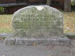 A stone gravestone with a curved top, with several lines of inscription. Trees, grass, and a wall are visible in the distance.