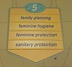 A yellow sign with a pointed bottom. At the top is the number 5 in an oval with a blue background. Below it are the words "family planning", "feminine hygiene", "feminine protection" and "sanitary protection"