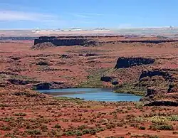 Reddish-brown terrain and many small green bushes surround a lake. Truncated ridges of dark rock run across the terrain parallel to the horizon and to each other beneath a blue sky.