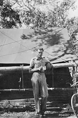Whitlam in military uniform stands under a tree in front of a large tent. He holds a mug in his hand.