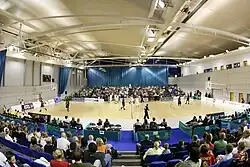 A Modern sporting facility. The building is roughly rectangular in shape and is quite modern, with a wooden clad design. It is surrounded by greenery and has several tall, white spikes on its roof.