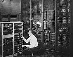 Black-and-white image of a man replacing one vacuum tube out of hundreds in an early computer