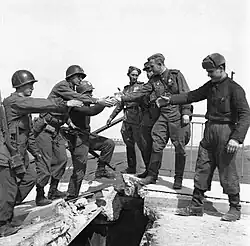 Black and white photo of a Soviet officer standing center-frame behind a handshake between a US and Soviet soldier on a wrecked bridge.