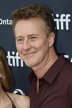 A portrait of Edward Norton, a blonde Caucasian man in a burgundy collared shirt. He is smiling to the left the camera.
