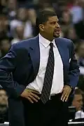 A man coaching from the sidelines of a basketball game. He is wearing a blue jacket and a white shirt with a dark, patterned tie.