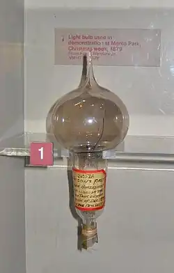 The inside of a museum exhibit shows a heavily used light-bulb with smoky glass. There is a handwritten inscription on a label wrapped around the stem of the bulb.