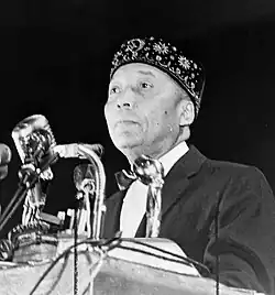 A clean-shaven, middle-aged black man, wearing an embroidered hat, standing at a podium with microphones in front of him