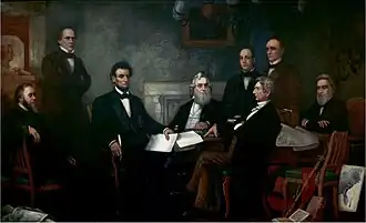 A dark-haired, bearded, middle-aged man holding documents is seated among seven other men.