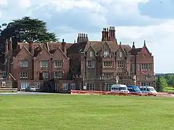 A large, three-storey house of red brick with stone dressings and tiled roof, with multiple bays, gables and chimneys, set in open grassed surroundings.