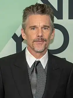 A Caucasian male with gray hair, wearing a two-piece dark gray suit with a gray and white shirt
