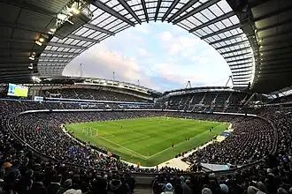 A football stadium as viewed from one corner. The photograph is a fish-eye paorama taken during a game against a blue sky. The stadium is full of fans on all four sides of the pitch, with large stands stretching up below an arched roof.