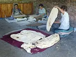 Three women make lavash: one prepares dough, one rolls it out, and one removes a lavash from the clay oven in the ground.