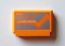 An orange plastic video game cartridge shaped in a flattened rectangular prism. The metallic light silver label has orange Japanese Katakana that translates to: "Family Computer. Mario Brothers." Below it is orange English text that reads "Mario Bros. © Nintendo 1983. Made in Japan."