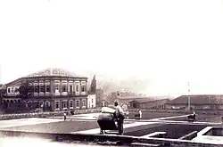 A photograph showing workers spreading or gathering coffee beans drying on a large paved plaza with an elegant, two-story neoclassical building on the left and warehouses and other plantation buildings in the background