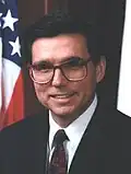 Hispanic man with large glasses and black hair with the US flag behind him