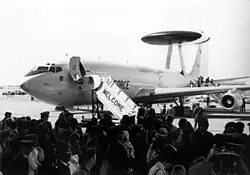 Black-and-white photograph with angled front view of four-engine jet aircraft on ramp with front fuselage door opened: A contingent of people are there to welcome the jet, which has a disc-shaped radar perching on top of struts on the dorsal fuselage.