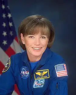 Formal portrait of astronaut in a flight suit displaying mission patches with an American flag in the background