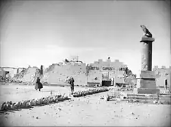 Two Allied soldiers, in greatcoats, walk towards a bullet-ridden and partially destroyed wall topped with crenelations. A bullet-riddled column stands in the foreground.