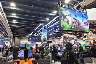 The Fortnite Battle Royale booth at GDC, featuring a large overhead screen on the right displaying the game's logo and demo stations on the left. People are dispersed around the booth.