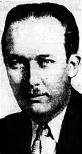Black-and-white portrait of an adult man, partly out of perspective, with slicked-back hair and a toothbrush mustache. He wears a suit.