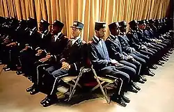 A group of black men, wearing navy blue uniforms, seated in two lines which intersect to form a V formation