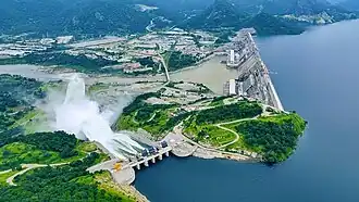 A photograph of large concrete dam and the reservoir it creates. The photo is taken from an aerial viewpoint, looking down from above. Large plumes of white water spray over a spillway.